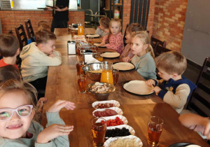 Zdjęcie przedstawia dzieci siedzące przy stole, czekające na podkład pizzy.