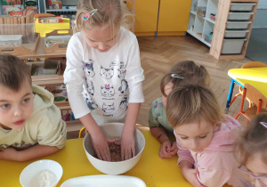 Zdjęcie przedstawia dzieci obserujące pracę dziewczynki podczas ugniatania ciasta.