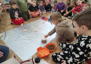 Zdjęcie przedstawia dzieci zgromadzone wokół sylwety choinki narysowanej na dużej folii. Jedno z dzieci wybiera drewnianą bombkę.