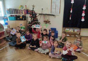 Zdjęcie przedstawia dzieci pozujące do zdjęcia. Dzieci siedzą zgromadzone na podłodze a w rękach trzymają świąteczne kartki. W tle stoi udekorowana choinka.
