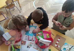 Zdjęcie przedstawia troje dzieci siedzących przy stole, na którym leżą materiały plastyczne. Dzieci robią kartki świąteczne.