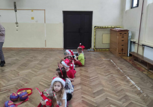 Zdjęcie przedstawia dzieci siedzące na podłodze w rzedzie na sali gimnastycznej. Dzieci mają w strojach mikołajkowe elementy.