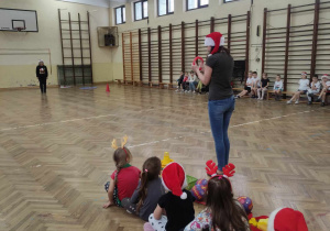 Zdjęcie przedstawia dzieci siedzące w rzędzie. Dzieci słuchają kobiety tłumaczącej im konkurencję.