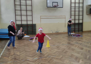 Zdjęcie przedstawia chłopca biorącego udział w konkurencji. Chłopiec biegnie przez salę gimnastyczną. W tle stoją nauczycielki.