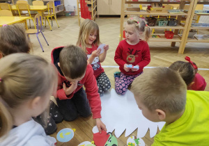 Zdjęcie przedstawia dzieci zgromadzone wokół białej papierowej choinki. Dzieci wybierają papierowe sylwety bombek.