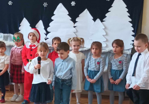Zdjęcie przedstawia dwoje dzieci mówiących do mikrofonu. Za nimi w rzędzie stoją inne dzieci. Wszyscy są ubrani odświętnie. W tle wisi zimowa dekoracja.