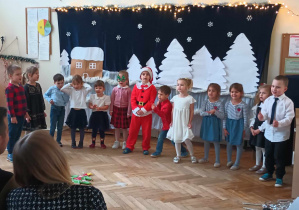 Zdjęcie przedstawia dzieci śpiewające piosenkę. Wszyscy są ubrani odświętnie. W tle wisi zimowa dekoracja.