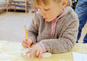 Zdjęcie przedstawia dziewczynkę siedzącą przy stole. Dziewczynka rysuje po drewnianej zabawce.