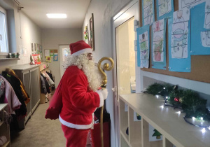 Zdjęcie przedstawia Mikołaja w przedszkolnym korytarzu. Mikołaj zagląda do jednej z sal.