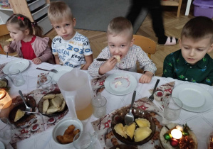 Na zdjęciu widzimy kilkoro dzieci siedzących przy stole wigilijnym. Jeden chłopiec je pieroga.