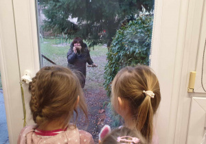 Na zdjęciu widzimy panią w ogródku przedszkolnym, która udaje, że obgryza ziarenka ptaszkom. Na panią patrzą przez okno dwie dziewczynki.