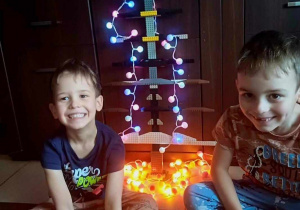 Zdjęcie przedstawia dwóch chłopców uśmiechających się do zdjęcia. Między nimi stoi choinka zbudowana z klocków ustrojona światełkami.