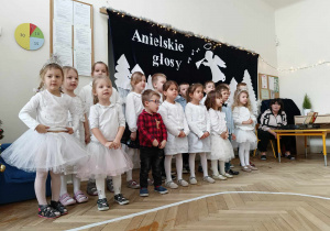 Zdjęcie przedstawia dzieci ustawione w dwóch rzędach i śpiewające piosenkę. W tle znajduje się biała dekoracja z napisem Anielskie granie.