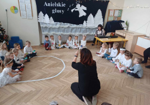 Zdjęcie przedstawia dzieci siedzące na podłodze, trzymające w rękach instrumenty. Przed dziećmi siedzi nauczycielka, która kieruje ich grą.