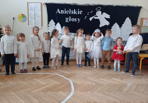 Zdjęcie przedstawia dzieci ustawione w rzędzie i śpiewające piosenkę. W tle znajduje się biała dekoracja z napisem Anielskie granie.