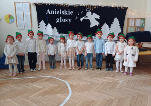 Zdjęcie przedstawia dzieci ustawione w rzędzie. Dzieci mają na głowach czapki elfów. W tle znajduje się biała dekoracja z napisem Anielskie granie.