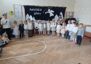 Zdjęcie przedstawia dzieci ustawione w rzędzie. W tle znajduje się biała dekoracja z napisem Anielskie granie.