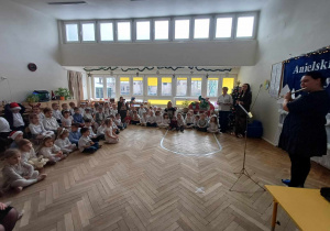 Zdjęcie przedstawia dzieci zgromadzone w sali, siedzące w półkolu. Przed nimi znajduje się nauczycielka grająca na instrumencie.