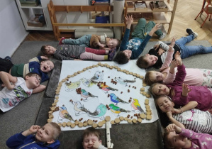 Na zdjęciu widzimy grupkę dzieci, która leży tworząc koło. Na środku na dywanie leży wykonany karmnik i ptaszki w nim.