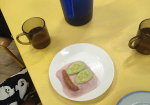 Zdjęcie przedstawia stół z talerzykami, kubkami i dzbankiem. Na jednym z talerzy leży kanapka, na której produkty spożywcze ułożone są w uśmiech.