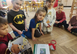 Zdjęcie przedstawia dzieci siedzące w kole. Na podłodze leży skoroszyt, stoi słoik z karteczkami w środku oraz torba z misiem uszatkiem.