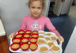 Na zdjęciu widać dziewczynkę, która na tacy zanosi kucharzowi babeczki marchewkowe do upieczenia