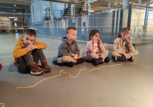 Zdjęcie przedstawia dzieci siedzące na podłodze. Przed dziećmi leżą rozciągnięte żółte sznurki.