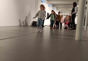 Zdjęcie przedstawia dzieci ustawione w rzędzie. Przed nimi rozłożony jest na podłodze sznur, po którym przechodzi jedna z dziewczynek.