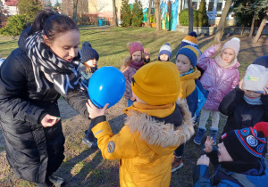 Zdjęcie przedstawia dzieci i nauczycielkę zgromadzonych w ogrodzie. Nauczycielka przekazuje jednemu z dzieci niebieski balonik.