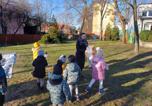 Zdjęcie przedstawia dzieci i nauczycielkę znajdujących się w ogrodzie. Wszyscy mają w rękach torebki foliowe, w które próbują złapać powietrze.