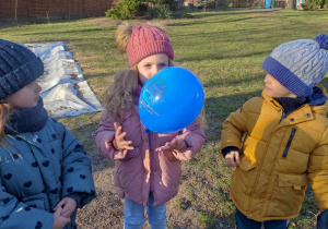 Zdjęcie przedstawia dzieci ustawione w ogrodzie. Dzieci przekazują sobie niebieski balonik.