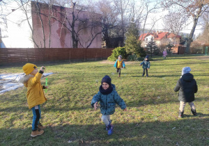Zdjęcie przedstawia dzieci biegające po ogrodzie. Jedno z nich puszcza bańki mydlane.