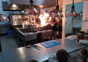 Na zdjęciu widzimy pana kucharza, który gotuje dla dzieci dania. Z patelni wydobywa się ogień.