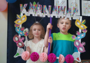 Zdjęcie przedstawia dwoje dzieci, które pozują do zdjęcia trzymając w rękach ramkę z napisem karnawał. Dzieci przebrane są w stroje.