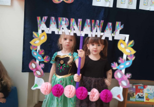 Zdjęcie przedstawia dwie dziewczynki, które pozują do zdjęcia trzymając w rękach ramkę z napisem karnawał. Dziewczynki przebrane są w stroje.