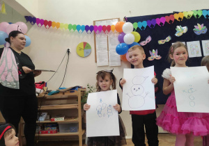 Zdjęcie przedstawia dzieci i nauczycielkę prowadzącą zabawę. Dzieci trzymają przed sobą kartki z narysowanymi bałwankami.