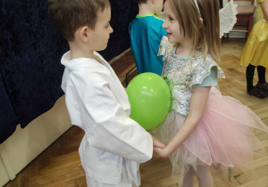 Zdjęcie przedstawia dwoje dzieci tańczących trzymając się za rączki. Między dziećmi znajduje się zielony balonik.