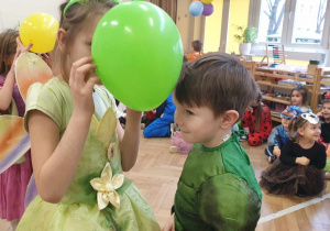 Na zdjęciu widzimy dziewczynkę i chłopca, którzy uczestniczą w zabawie karnawałowej. Trzymają pomiędzy swoimi czołami zielonego balona i tańczą do piosenki.