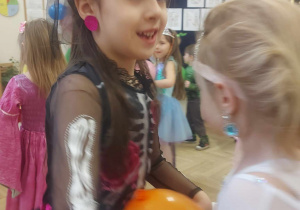 Na zdjęciu widzimy dwie dziewczynki, które uczestniczą w zabawie. Pomiędzy soimi brzuchami trzymają pomarańczowego balona.