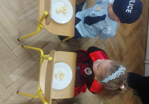 Na zdjęciu widzimy dzieci, które uczestniczą w zabawie. Na krzesełkach leżą talerze, na których znadjują się pokrojone jabłka. Zadaniem dzieci było zjedzenie ich bez użycia rąk.