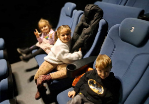 Zdjęcie przedstawia dzieci siedzące na fotelach w sali kinowej.