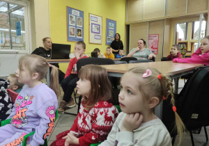 Zdjęcie przedstawia dzieci patrzące przed siebie. Część dzieci siedzi przy stołach. Na zdjęciu znajdują się również trzy kobiety, w tym prowadząca zajęcia.