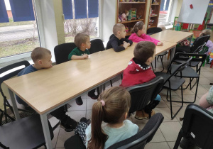 Zdjęcie przedstawia dzieci siedzące przy stole. Dzieci słuchają osoby prowadzącej zajęcia.