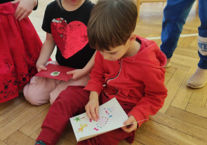 Zdjęcie przedstawia chłopca i dziewczynkę, którzy siedzą na podłodze i przyglądają się kartce, którą chłopiec trzyma w ręku.