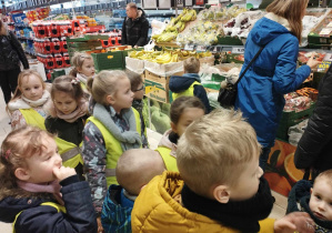 Na zdjęciu widzimy grupkę dzieci, które stoją przy półkach sklepowych i szukają produktów ekologicznych.