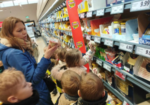 Na zdjęciu widzimy dzieci, które stoją przy sklepowych lodówkach i szukają produktów z kuchni włoskiej, francuskiej i produktów ekologicznych. W gronie tym stoi także nauczyciel, który trzyma w rękach bio jogurt.