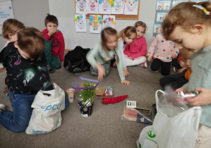 Na zdjęciu widzimy dzieci siedzące na dywanie w klasie. Dwójka z nich wyjmuje zakupione produkty z toreb.