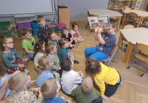 Zdjęcie przedstawia chłopca pokazującego ilustracje w książce innym dzieciom.