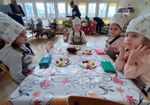 Dzieci siedzą przy stole i czekają na foremki z płynnną czekoladą.