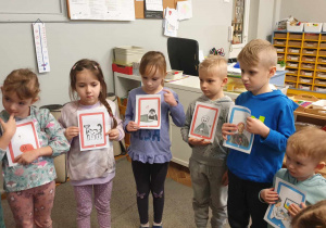 Zdjecie przedstawia stojące dzieci, trzymające różne ilustracje w czerwonych ramkach.